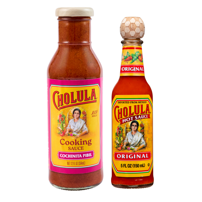 A bottle of Cholula Cooking Sauce in Cochinita Pibil flavor stands next to a bottle of Cholula Original Hot Sauce against a black background.