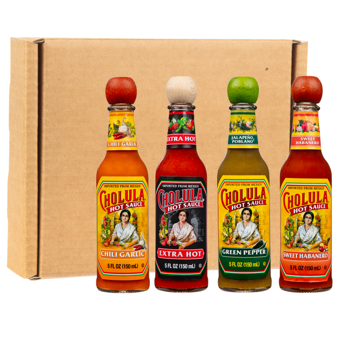 Four bottles of Cholula Hot Sauce, a McCormick brand, are arranged in a row in front of a cardboard box, showcasing the Chili Garlic, Extra Hot, Green Pepper, and Sweet Habanero flavors.