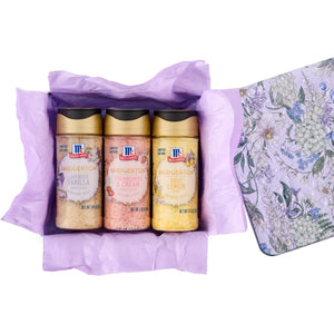 A gift set featuring McCormick Bridgerton Limited Edition Finishing Sugars in Lavender Vanilla, Strawberries &amp; Cream, and Sweet Lemon flavors is displayed in a floral-patterned tin with purple tissue paper.