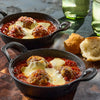 Two individual cast-iron skillets are filled with large meatballs in a rich tomato sauce, topped with melted mozzarella cheese, and served with a side of crusty bread.