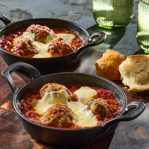 Two individual cast-iron skillets are filled with large meatballs in a rich tomato sauce, topped with melted mozzarella cheese, and served with a side of crusty bread.