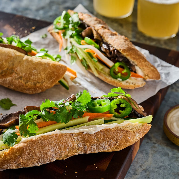 Two mushroom banh mi sandwiches are filled with fresh cilantro, jalapeños, and pickled vegetables, served on a wooden board with two glasses of beer in the background.