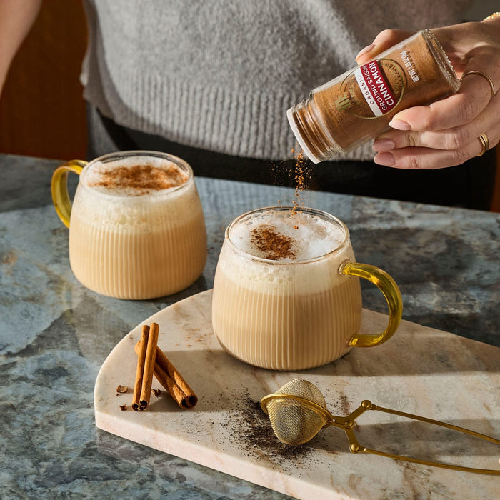 A person sprinkles McCormick Organic Ground Saigon Cinnamon from a glass jar onto the foam of a warm beverage in a ribbed glass mug.