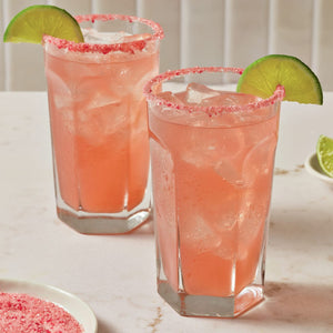 Two tall glasses of a pink iced beverage are garnished with a lime wedge and a rim of Mccormick Strawberry Finishing Sugar, 3.67 Oz.