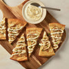 An overhead shot shows several slices of flatbread pizza on a wooden board, garnished with a creamy drizzle, green onions, and a sprinkle of Mccormick Ranch Finishing Salt, 4.03 Oz, next to a bowl of seasoned dip.