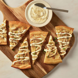 An overhead shot shows several slices of flatbread pizza on a wooden board, garnished with a creamy drizzle, green onions, and a sprinkle of Mccormick Ranch Finishing Salt, 4.03 Oz, next to a bowl of seasoned dip.