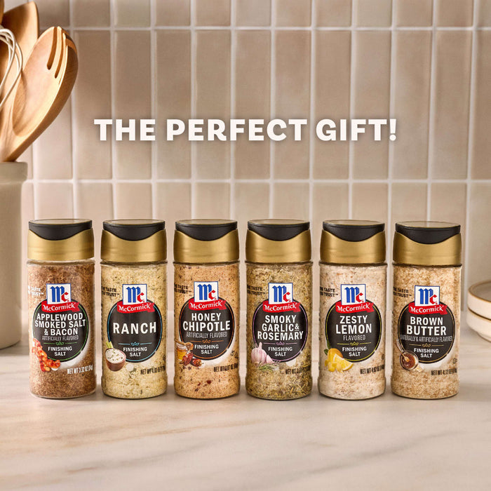 A lineup of six different McCormick Finishing Salts, including Applewood Smoked Salt &amp; Bacon and Zesty Lemon, are displayed on a kitchen counter with text above that reads, The Perfect Gift!