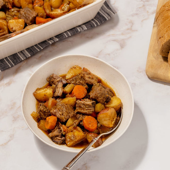 A hearty bowl of beef stew featuring tender meat and vegetables prepared using the Dtc Bundle sits on a marble countertop next to a loaf of sliced bread.