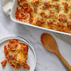 A serving of cheesy baked rotini pasta from the Dtc Bundle sits on a white plate next to the full casserole dish and a wooden spoon.