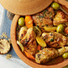 A close-up of a tagine filled with chicken, olives, and vegetables, with a small bowl of Ducros Coriander From France 7G spice next to it.
