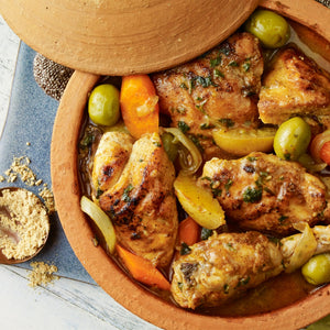 A close-up of a tagine filled with chicken, olives, and vegetables, with a small bowl of Ducros Coriander From France 7G spice next to it.