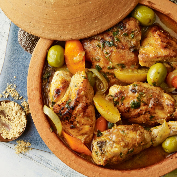 A close-up of a tagine filled with chicken, olives, and vegetables, with a small bowl of Ducros Coriander From France 7G spice next to it.