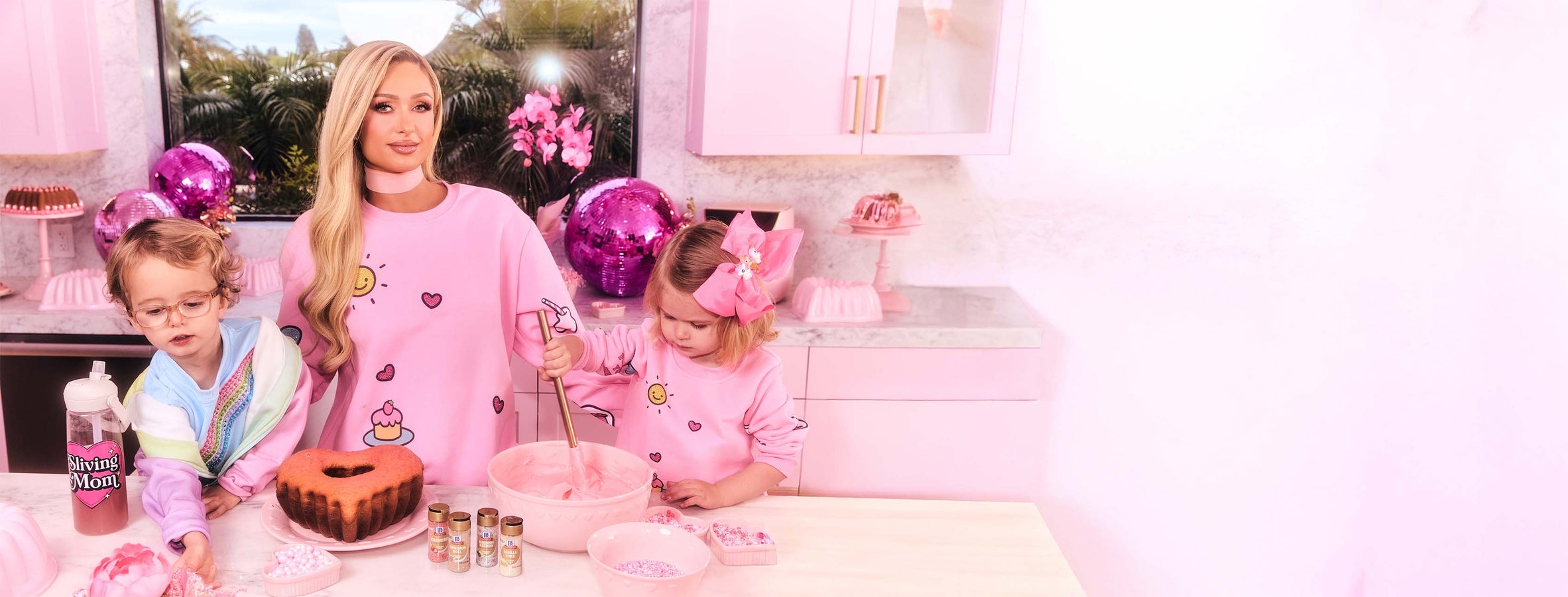 Paris Hilton and two children in a kitchen with pink decor and balloons.