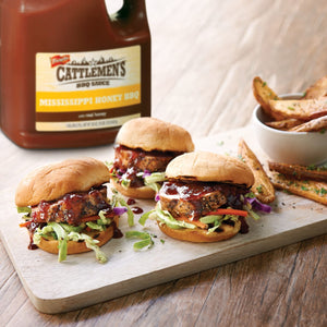 Three mini burgers topped with coleslaw and Cattlemens BBQ sauce on a wooden cutting board with potato wedges in the background.