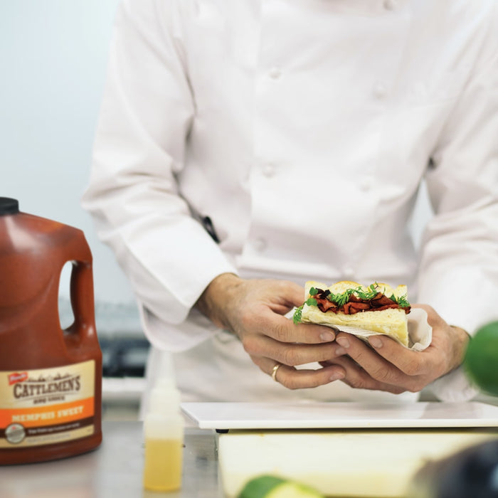 A person in a white chefs coat holds a sandwich in their hands with a bottle of Cattlemens Memphis Sweet BBQ Sauce in the background.