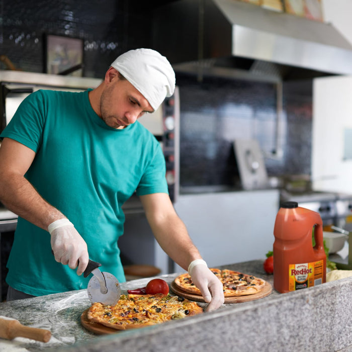A man in a white hat is cutting a pizza with a pizza cutter while wearing white gloves and a green shirt with a bottle of Franks RedHot sauce in the background.