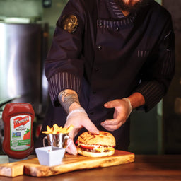 A chef wearing gloves is placing a burger on a wooden cutting board next to a bottle of Frenchs ketchup.