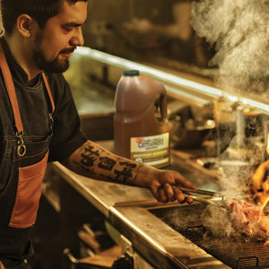 A chef is grilling a steak on a grill with a bottle of Cattlemens Kentucky Bourbon in the background.