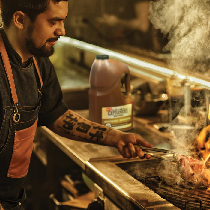A chef is grilling a steak on a grill with a bottle of Cattlemens Kentucky Bourbon in the background.