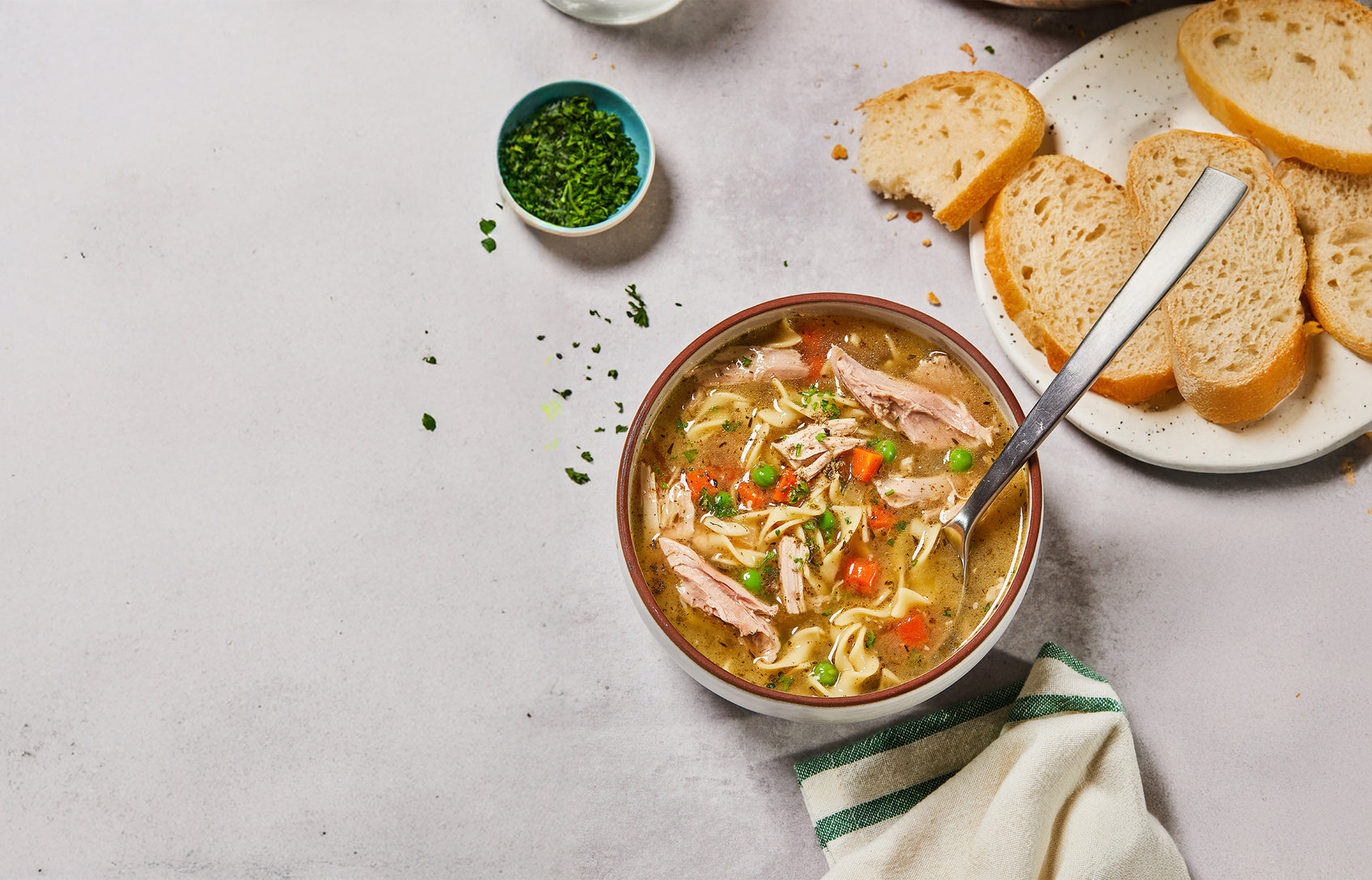 Bowl of soup with noodles, carrots, and peas on a table with bread slices and a small bowl of greens.