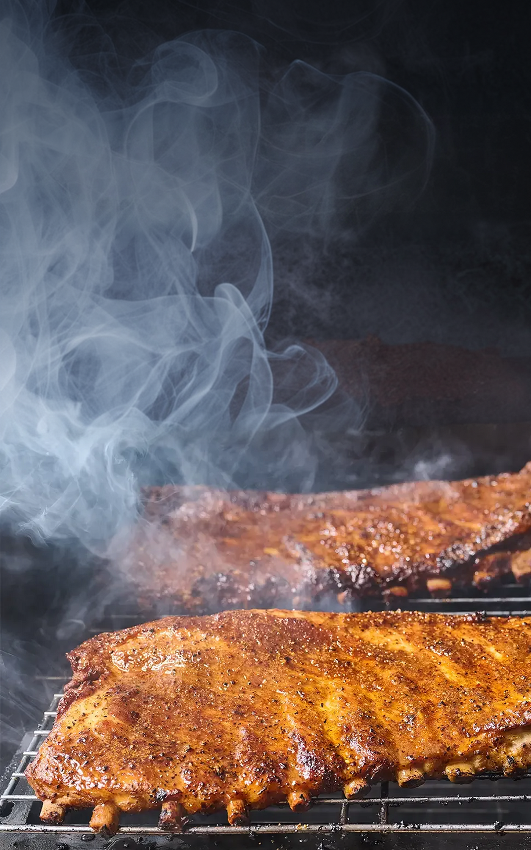 Spiced ribs cooking on a grill with smoke rising against a dark background
