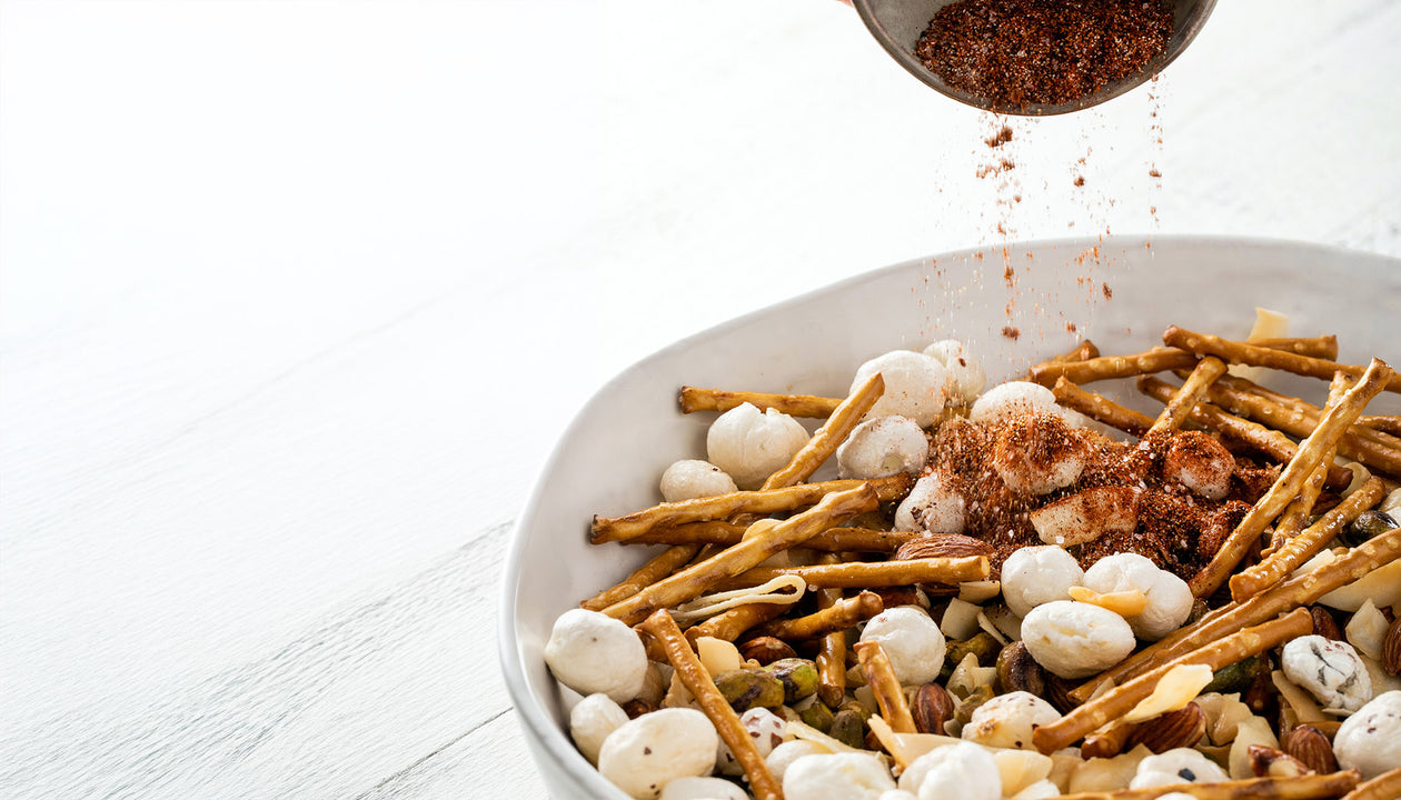Spice blend being sprinkled over a bowl of snack mix with pretzel sticks, nuts, and puffed crackers on a white surface.