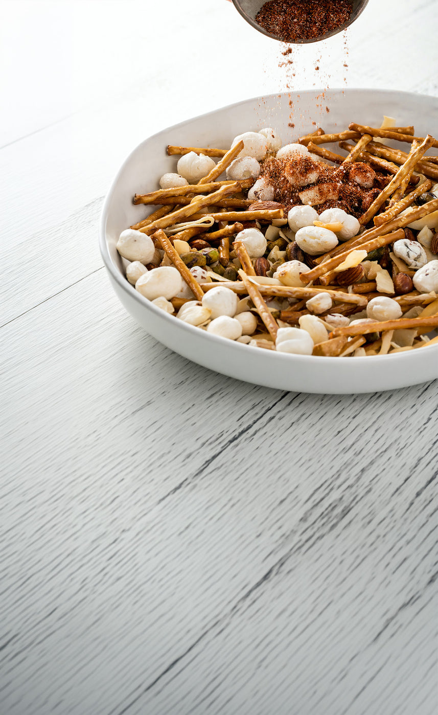 Spice blend being sprinkled over a bowl of snack mix with pretzel sticks, nuts, and puffed crackers on a white surface.