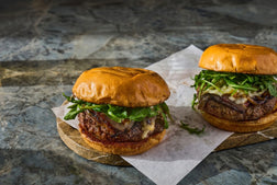 Two gourmet beef burgers with fresh arugula, melted cheese, and crispy fried onions on toasted buns, served on a wooden board with parchment paper on a dark marble surface.