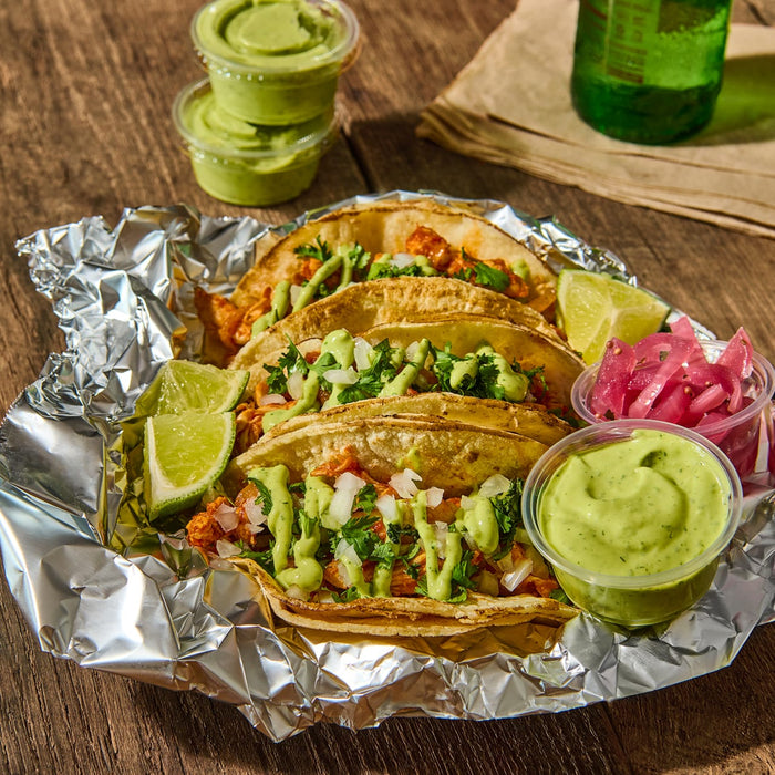 Three tacos with cilantro onions and avocado crema on a bed of aluminum foil with lime wedges pickled onions and a container of avocado crema.