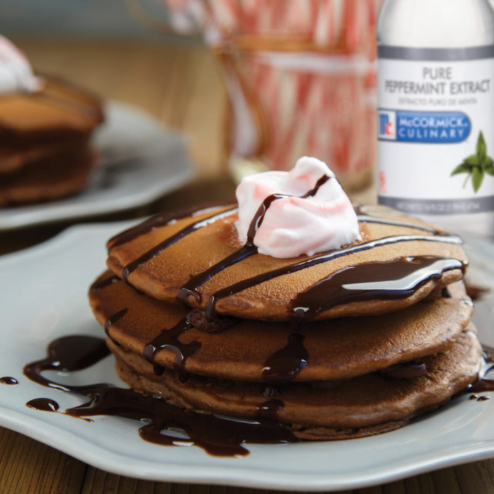 A plate of chocolate pancakes topped with whipped cream and chocolate sauce, with a bottle of McCormick Culinary Pure Peppermint Extract in the background.
