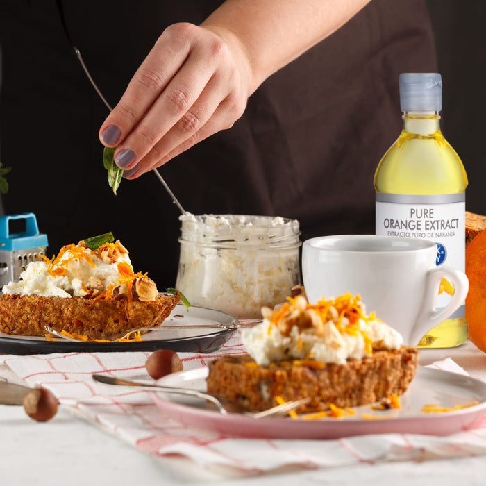 A hand is adding a sprig of mint to a plate of carrot cake topped with cream cheese, orange zest, and hazelnuts.