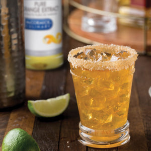 A glass of Pure Peach Coconut cocktail with ice and a sugared rim, next to a lime wedge and a bottle of Pure Peach Coconut.