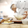 A chef in a white uniform is mixing batter in a glass bowl with a whisk, using McCormick Culinary Imitation Almond Extract.