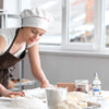A baker in a white chefs hat rolls out dough on a floured surface with a rolling pin.