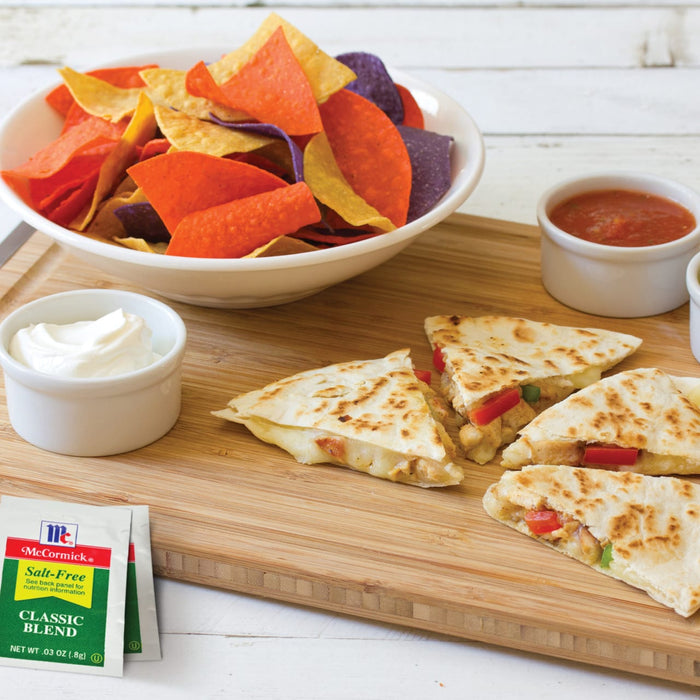 A close-up of a wooden cutting board with a bowl of tortilla chips, a bowl of sour cream, a bowl of salsa, and three quesadillas with a packet of McCormick Salt-Free Classic Blend seasoning.