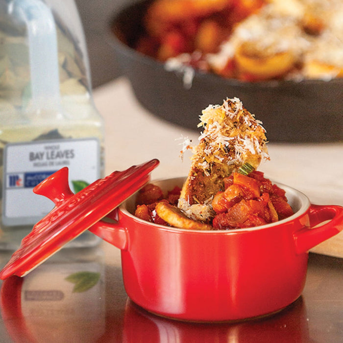 A red Le Creuset pot with a lid on top, filled with a tomato-based dish topped with a piece of toasted bread.