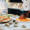 A wooden spoon is spreading tomato sauce on a pizza crust, with a finished pizza and a jar of McCormick Culinary Garlic Powder in the background.