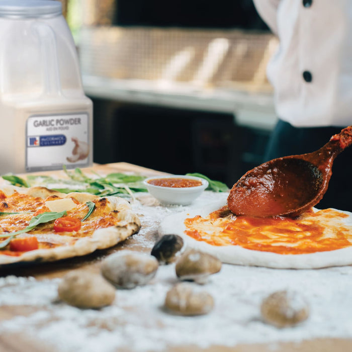 A wooden spoon is spreading tomato sauce on a pizza crust, with a finished pizza and a jar of McCormick Culinary Garlic Powder in the background.