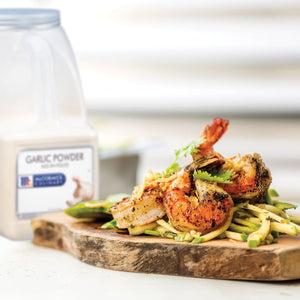 A wooden cutting board with a serving of grilled shrimp with zucchini noodles and a container of McCormick Garlic Powder.