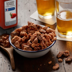 A bowl of McCormick Culinary Smoked Paprika Cashews and Pecans with two glasses of beer in the background.