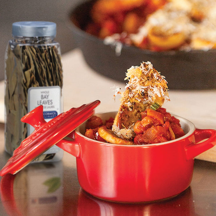 A red pot with a lid filled with a tomato based dish topped with toasted bread and parmesan cheese next to a jar of McCormick Whole Bay Leaves.