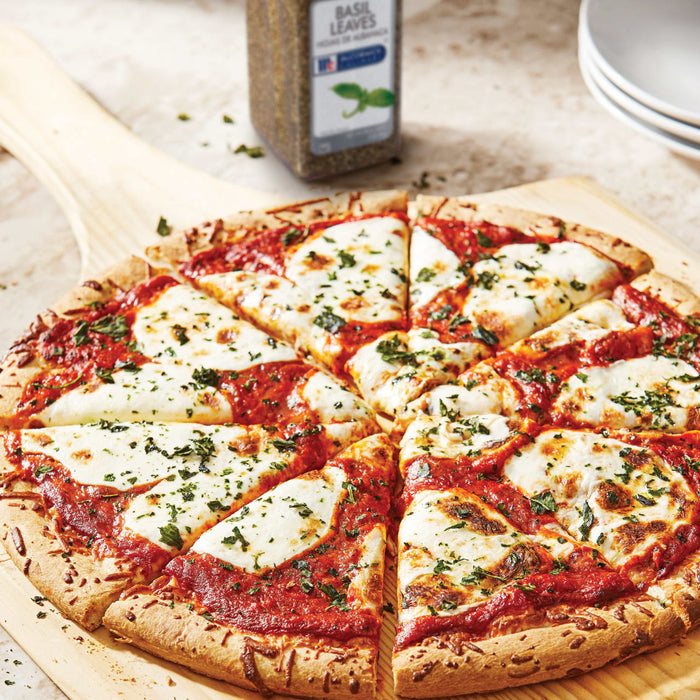 A pizza with mozzarella cheese and tomato sauce on a wooden cutting board with a jar of McCormick Basil Leaves.