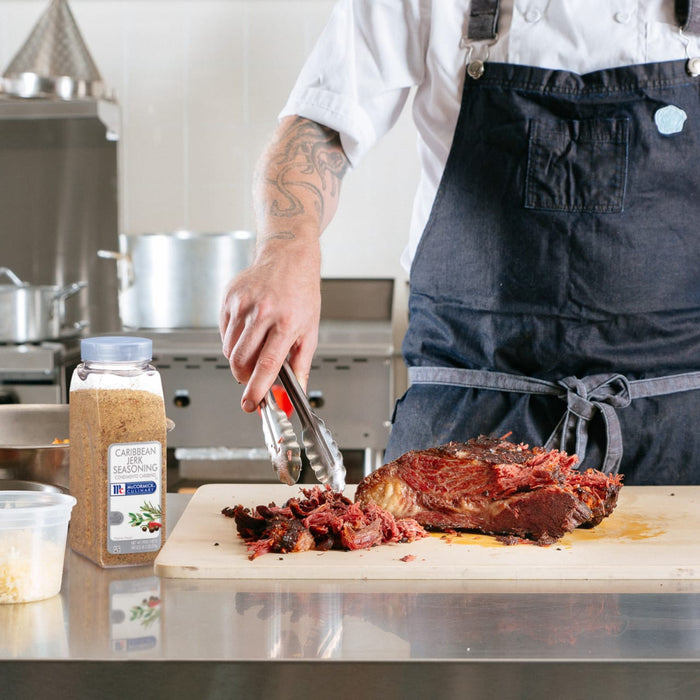A chef is using tongs to pull apart a piece of jerk seasoned meat on a cutting board next to a container of McCormick Caribbean Jerk Seasoning.