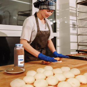A baker wearing blue gloves rolls out dough on a wooden counter with a McCormick Ground Cinnamon container in the background.