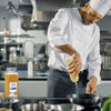 A chef in a white uniform and black and white striped apron pours oil into a pan on a stovetop next to a jar of McCormick Culinary Curry Powder.