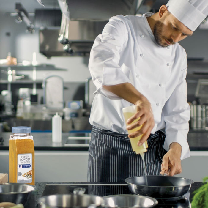 A chef in a white uniform and black and white striped apron pours oil into a pan on a stovetop next to a jar of McCormick Culinary Curry Powder.