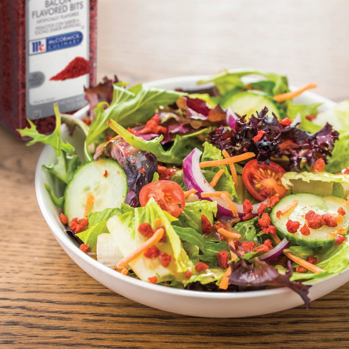 A bowl of salad with bacon bits cucumbers tomatoes and red onion with a jar of McCormick Culinary Bacon Flavored Bits in the background.
