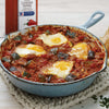 A blue cast iron skillet with a handle sits on a white crocheted placemat and contains a dish of shakshuka with four eggs zucchini and tomatoes.
