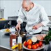 A chef in a white uniform is preparing food in a kitchen using tongs to stir a pan of food.
