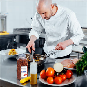 A chef in a white uniform is preparing food in a kitchen using tongs to stir a pan of food.